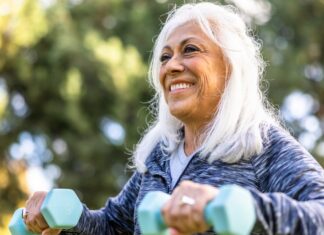 3 Anti-Aging Exercises That Will Keep You Fit as You Age An older adult with shoulder-length white hair and a blue patterned sweatshirt lifting light blue weights while surrounded by trees outside.