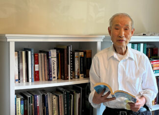 Home Improvements Can Help People Age Independently. But Medicare Seldom Picks Up the Bill. A senior man stands beside a bookshelf, holding a book. He wears a white button-shirt.